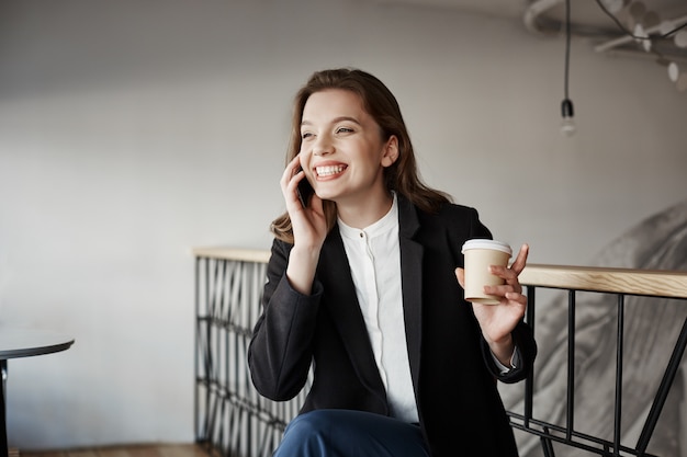 Retrato de hermosa mujer europea inteligente sentado en la cafetería, tomando café y gesticulando mientras habla por teléfono inteligente