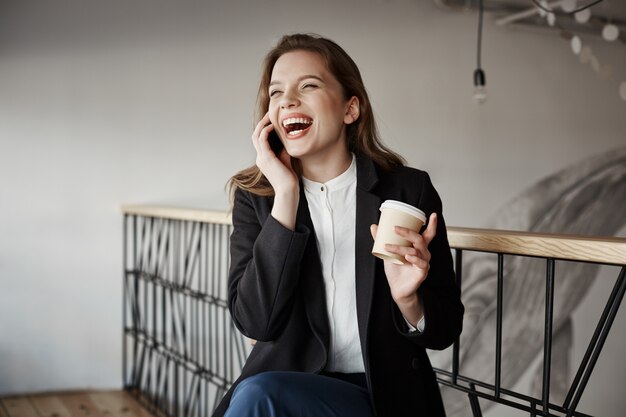 Retrato de hermosa mujer europea inteligente sentado en la cafetería, tomando café y gesticulando mientras habla por teléfono inteligente