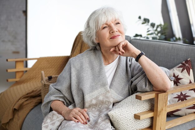 Retrato de hermosa mujer europea de pelo gris de mediana edad con sonrisa de ensueño y ojos llenos de sabiduría relajándose solo en casa, sentada en un cómodo sofá, recordando los días de su juventud