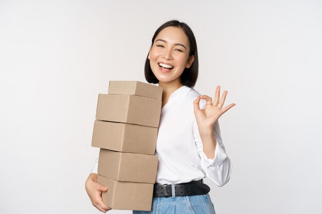 Retrato de hermosa mujer feliz llevar cajas con pedidos Empresaria con entrega de acarreo y mostrando bien signo fondo blanco