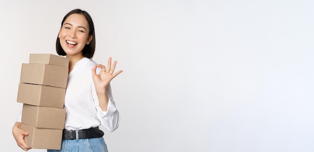 Retrato de hermosa mujer feliz llevar cajas con pedidos Empresaria con entrega de acarreo y mostrando bien signo fondo blanco