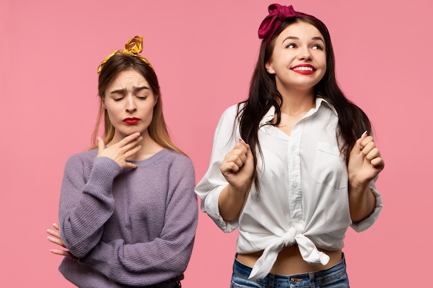 Retrato de hermosa mujer joven con cabello negro mirando hacia arriba con amplia sonrisa feliz, emocionada con noticias positivas, amiga disgustada de pie junto a ella con mirada escéptica dudosa