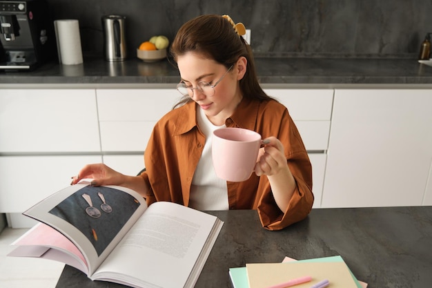 Foto gratuita retrato de una hermosa mujer joven en la cocina sentada con un libro volviendo páginas leyendo y bebiendo