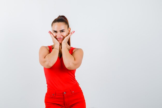 Retrato de hermosa mujer joven cogidos de la mano en las mejillas en camiseta roja, pantalones y mirando alegre vista frontal