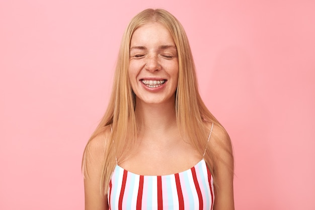 Retrato de hermosa mujer joven con pecas, perforaciones faciales y aparatos ortopédicos sonriendo con entusiasmo, manteniendo los ojos cerrados, esperando sorpresa