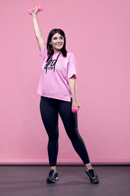 Retrato de una hermosa mujer latina positiva del cuerpo en una sudadera deportiva rosa con ejercicio con pesas en rosa