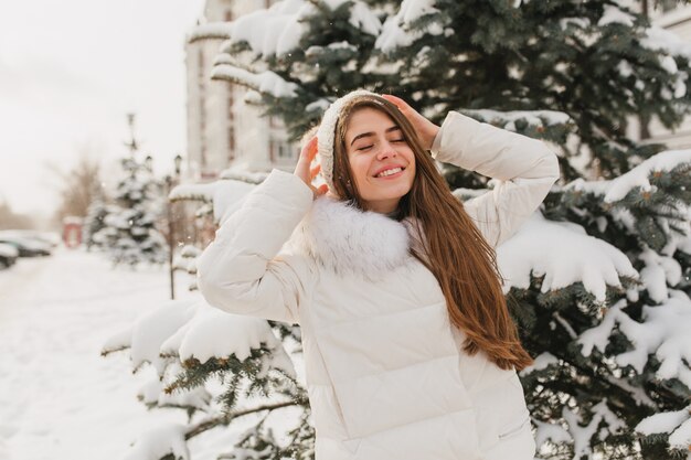 Retrato hermosa mujer linda escalofriante en el sol en la mañana helada. Mujer alegre joven que disfruta del invierno en abetos llenos de nieve. Emociones verdaderas positivas, sonriendo con los ojos cerrados.