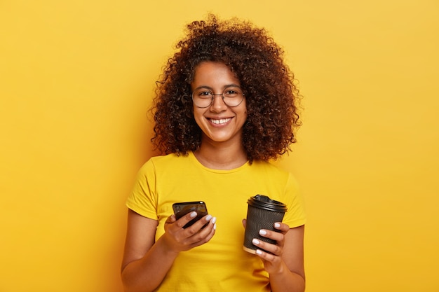 Retrato de hermosa mujer milenaria sostiene un teléfono inteligente y bebe café para llevar, sonríe agradablemente, usa una camiseta amarilla, pregunta el número para reunirse más tarde