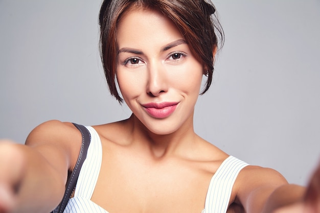 Retrato de hermosa mujer morena linda modelo en vestido casual de verano sin maquillaje haciendo foto selfie en teléfono aislado en gris con bolso