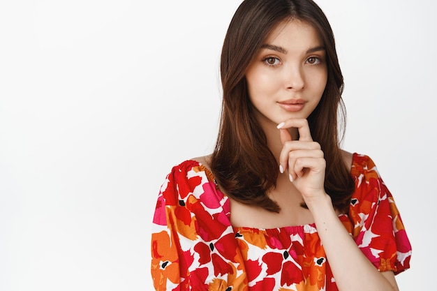 Retrato de una hermosa mujer morena mirando a la cámara pensativa usando un elegante vestido floral de pie sobre fondo blanco