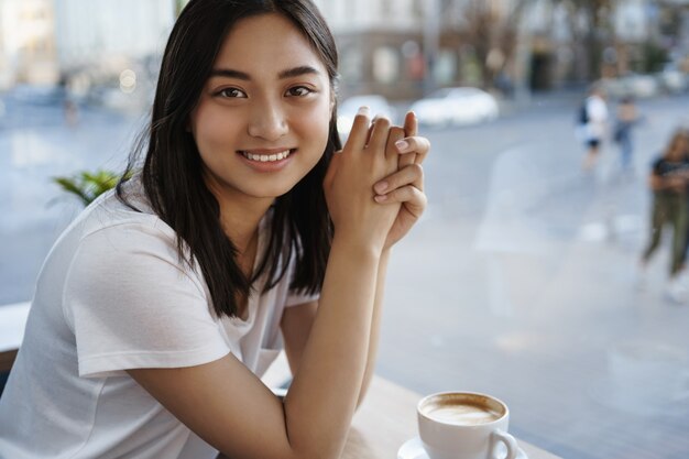Retrato de hermosa mujer natural tomando café en la cafetería solo, sentado junto a la ventana, sonriendo a la cámara feliz.