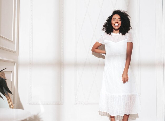 Retrato de hermosa mujer negra con peinado de rizos afro Modelo sonriente vestida con vestido blanco de verano Sexy mujer despreocupada posando cerca de la pared en el estudio Bronceada y alegre En un día soleado