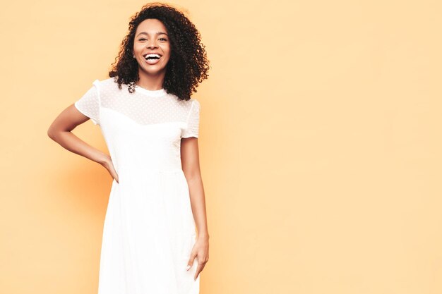 Retrato de hermosa mujer negra con peinado de rizos afro Modelo sonriente vestida con vestido blanco de verano Sexy mujer despreocupada posando junto a la pared amarilla en el estudio Bronceada y alegre Aislada