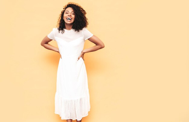 Retrato de hermosa mujer negra con peinado de rizos afro Modelo sonriente vestida con vestido blanco de verano Sexy mujer despreocupada posando junto a la pared amarilla en el estudio Bronceada y alegre Aislada