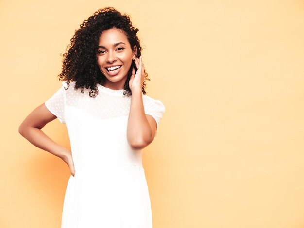 Retrato de hermosa mujer negra con peinado de rizos afro Modelo sonriente vestida con vestido blanco de verano Sexy mujer despreocupada posando junto a la pared amarilla en el estudio Bronceada y alegre Aislada