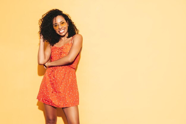 Retrato de hermosa mujer negra con peinado de rizos afro Modelo sonriente vestida con vestido rojo de verano Mujer sexy despreocupada posando junto a la pared amarilla en el estudio Bronceada y alegre Aislada