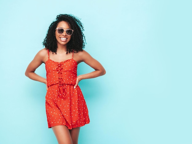 Retrato de hermosa mujer negra con peinado de rizos afro Modelo sonriente vestida con vestido rojo de verano Mujer sexy despreocupada posando junto a la pared azul en el estudio Bronceada y alegre En gafas de sol