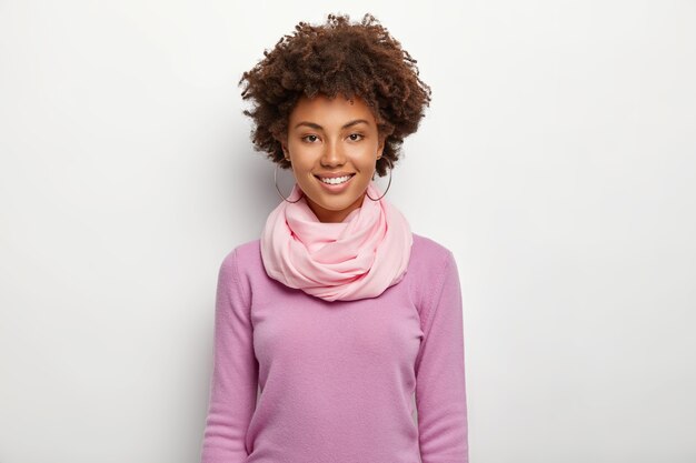 Retrato de hermosa mujer de pelo rizado viste jersey violeta púrpura y pañuelo de seda, mira directamente, expresa buenas emociones, modelos contra la pared blanca. Gente, etnia, moda