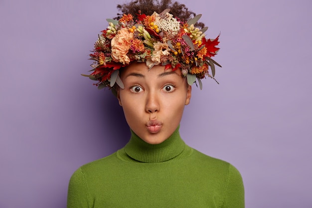 Foto gratuita el retrato de una hermosa mujer rizada mantiene los labios redondeados, tiene la piel oscura y saludable, usa una corona de otoño, hace muecas, vestida con un cuello alto verde.