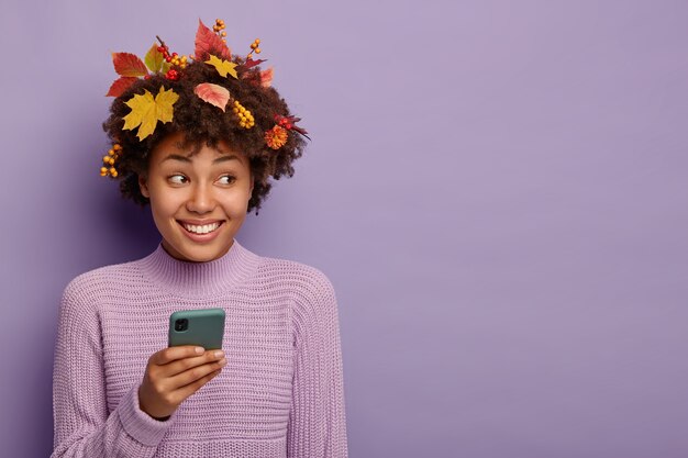 Retrato de hermosa mujer rizada usa teléfono inteligente, tiene hojas de otoño en la cabeza, está en alto espíritu, posa sobre fondo violeta