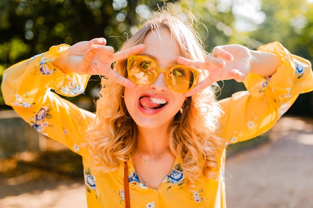 Retrato de hermosa mujer rubia con estilo emocional en blusa amarilla con gafas de sol, expresión divertida cara loca