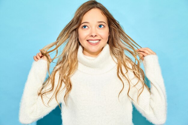 Retrato de hermosa mujer rubia hermosa sonriente. Mujer de pie en el elegante suéter blanco, en la pared azul. Concepto de invierno