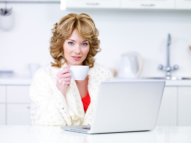 Retrato de hermosa mujer rubia con taza de café y portátil sentado en la cocina