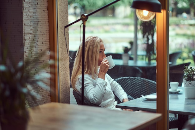 Retrato de una hermosa mujer rubia vestida con una blusa blanca sentada en una mesa bebe café en el café de la terraza.