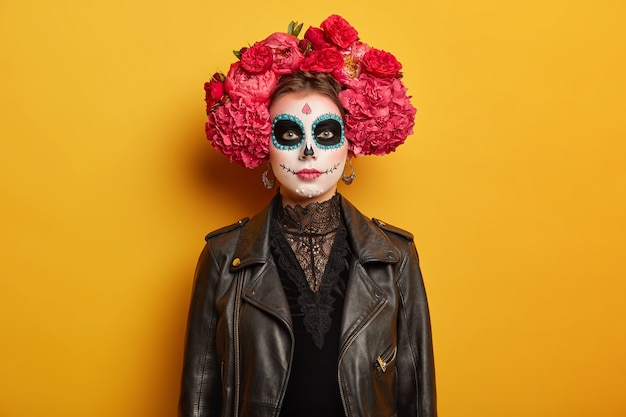 El retrato de una hermosa mujer seria tiene un maquillaje vívido creativo, usa una corona de flores, ropa negra, intenta dar miedo, viene en la fiesta de Halloween o el Día de los Muertos