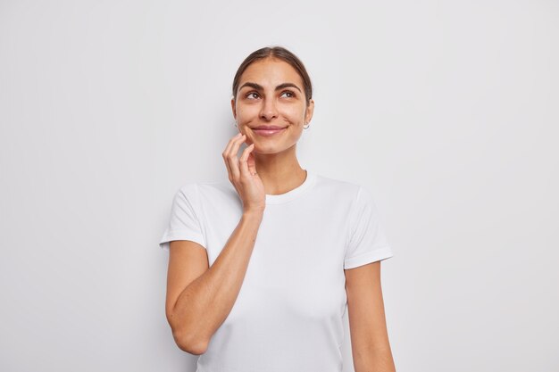 Retrato de hermosa mujer soñadora con cabello oscuro sonríe suavemente mira arriba recuerda agradables recuerdos vestida con camiseta casual aislada sobre pared blanca tiene pensamientos románticos se encuentra en el interior