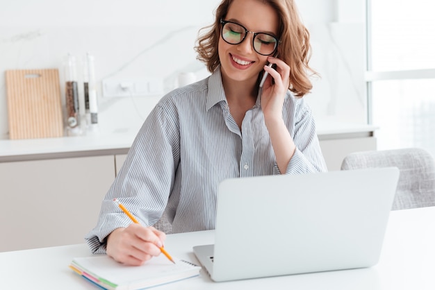 Retrato de hermosa mujer sonriente en gafas tomando notas mientras habla por teléfono móvil, en el interior