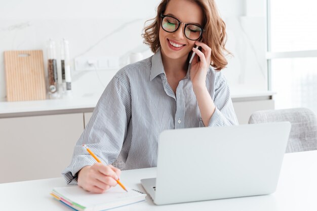 Retrato de hermosa mujer sonriente en gafas tomando notas mientras habla por teléfono móvil, en el interior