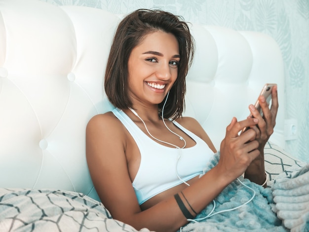 Retrato de hermosa mujer sonriente vestida con pijama blanco y escuchando música con auriculares