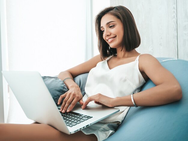 Retrato de hermosa mujer sonriente vestida con pijama blanco. Modelo despreocupado sentado en una silla de bolsa blanda y usando una computadora portátil.