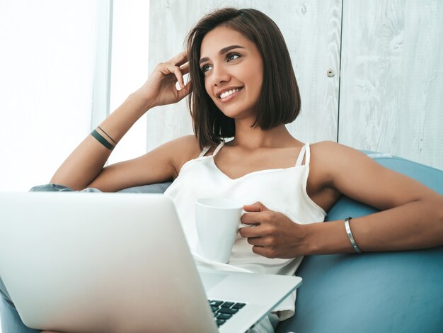 Retrato de hermosa mujer sonriente vestida con pijama blanco. Modelo despreocupado sentado en una silla de bolsa blanda y usando una computadora portátil.