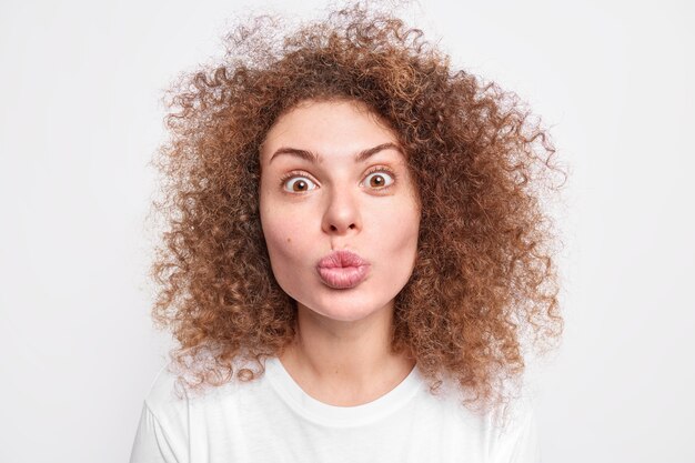 El retrato de la hermosa mujer sorprendida de pelo rizado hace pucheros con los labios que van a tener el primer beso en su vida envía miradas de mwah sorprendentemente vestidas casualmente aisladas sobre una pared blanca. Expresiones de la cara