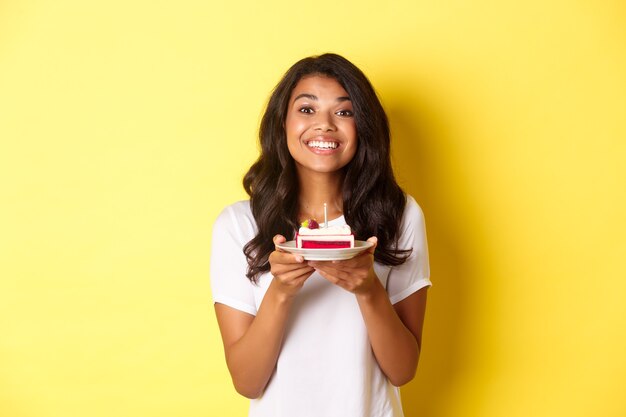 Retrato de hermosa niña afroamericana celebrando un cumpleaños, sonriendo y mirando feliz y sosteniendo b-day cake con vela, de pie sobre fondo amarillo