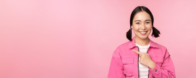 Retrato de una hermosa niña asiática sonriente señalando con el dedo a la izquierda mostrando una promoción de banner publicitario de pie sobre un fondo rosa