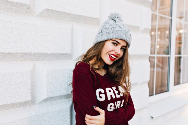 Retrato hermosa niña con cabello largo en suéter marsala en la pared gris exterior. Lleva gorro de punto, labios rojos y sonriente.