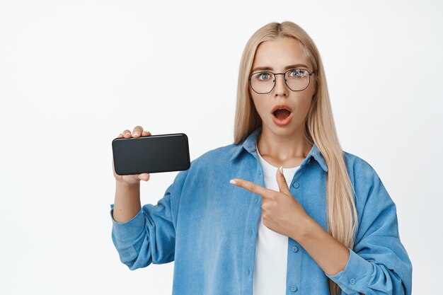 Retrato de hermosa niña mirando sorprendida, mostrando algo perturbador en la pantalla del teléfono móvil, apuntando al teléfono inteligente, de pie en blanco.