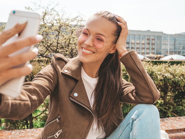 Retrato de hermosa niña morena sonriente en jeans y chaqueta hipster de verano Modelo tomando selfie en smartphone