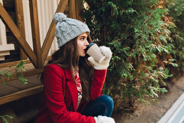 Retrato hermosa niña con pelo largo en abrigo rojo, gorro de punto y guantes blancos sentado en escaleras de madera cerca de ramas verdes al aire libre. Ella bebe café y mira a un lado.