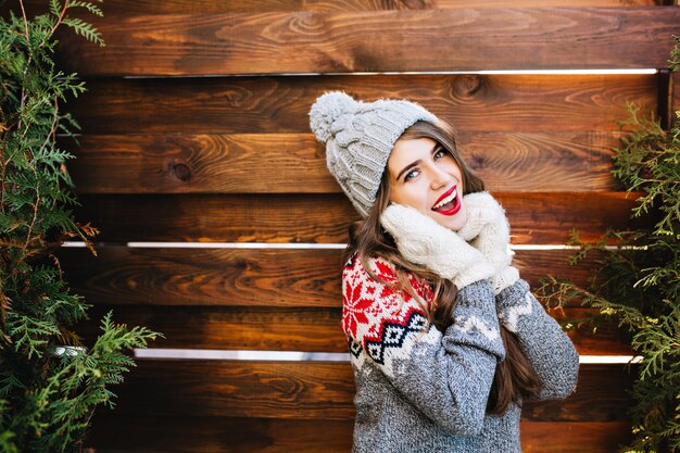 Retrato hermosa niña con pelo largo en sombrero de punto y suéter de invierno en madera. Ella toca la cara con las manos en guantes y sonríe.