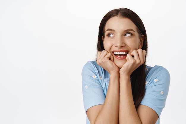 Foto gratuita retrato de una hermosa niña romántica, pensando en algo increíble, imaginando algo interesante, sosteniendo la cabeza en las manos, mirando a un lado con una sonrisa divertida en blanco.