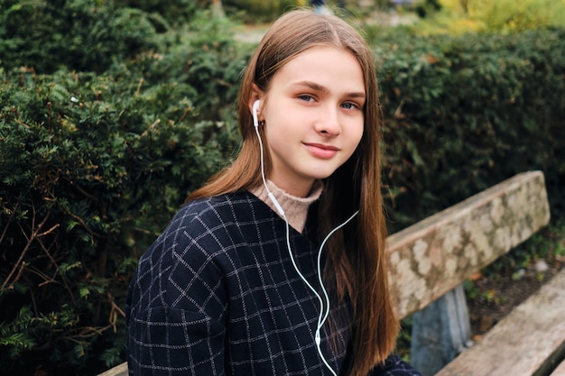 Retrato de una hermosa niña sonriente con auriculares mirando felizmente a la cámara en un banco en el parque de la ciudad