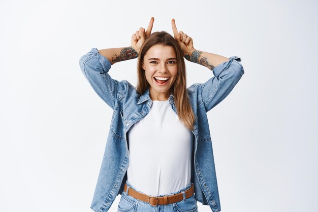 Retrato de una hermosa niña sonriente divirtiéndose mostrando lindos cuernos de dedos en la cabeza y luciendo feliz haciendo el tonto y jugando de pie sobre fondo blanco