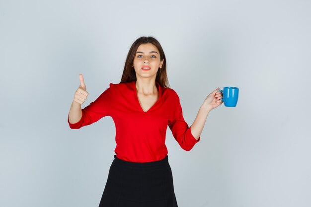 Retrato de hermosa niña sosteniendo la taza, mostrando el pulgar hacia arriba en blusa roja, falda negra y mirando pensativo, vista frontal.