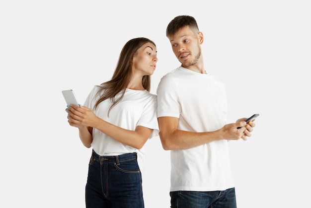 Retrato de hermosa pareja joven aislado sobre fondo blanco de estudio. Expresión facial, emociones humanas, concepto publicitario. Ambos sosteniendo teléfonos inteligentes, mujer mirando en su pantalla. Guardar secretos.