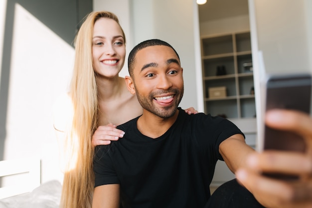 Retrato de hermosa pareja de linda joven sincera con cabello largo y rubio y chico guapo haciendo selfie en cama en apartamento moderno. Divirtiéndose, familia joven, sonriendo, felicidad