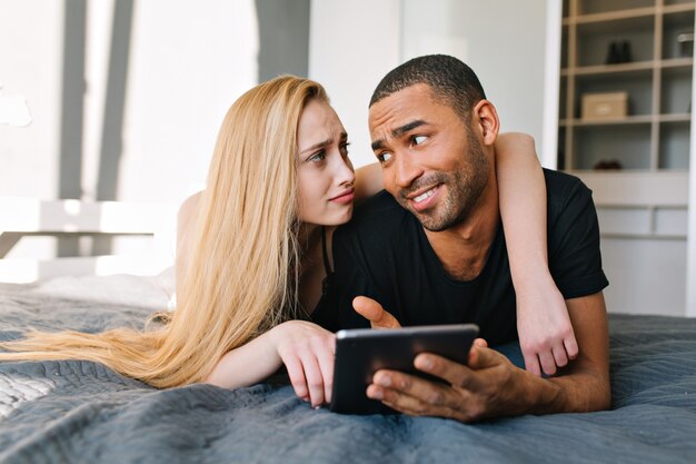 Retrato hermosa pareja de linda joven sincera con largo cabello rubio y chico guapo hablando en la cama en el apartamento moderno. Buscando en internet, divertirse, familia joven